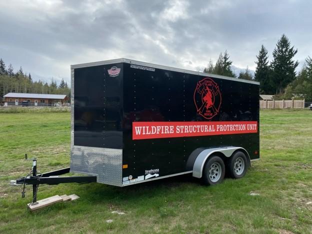 Black trailer with "WILDFIRE STRUCTURAL PROTECTION UNIT" text, set in a grassy area.