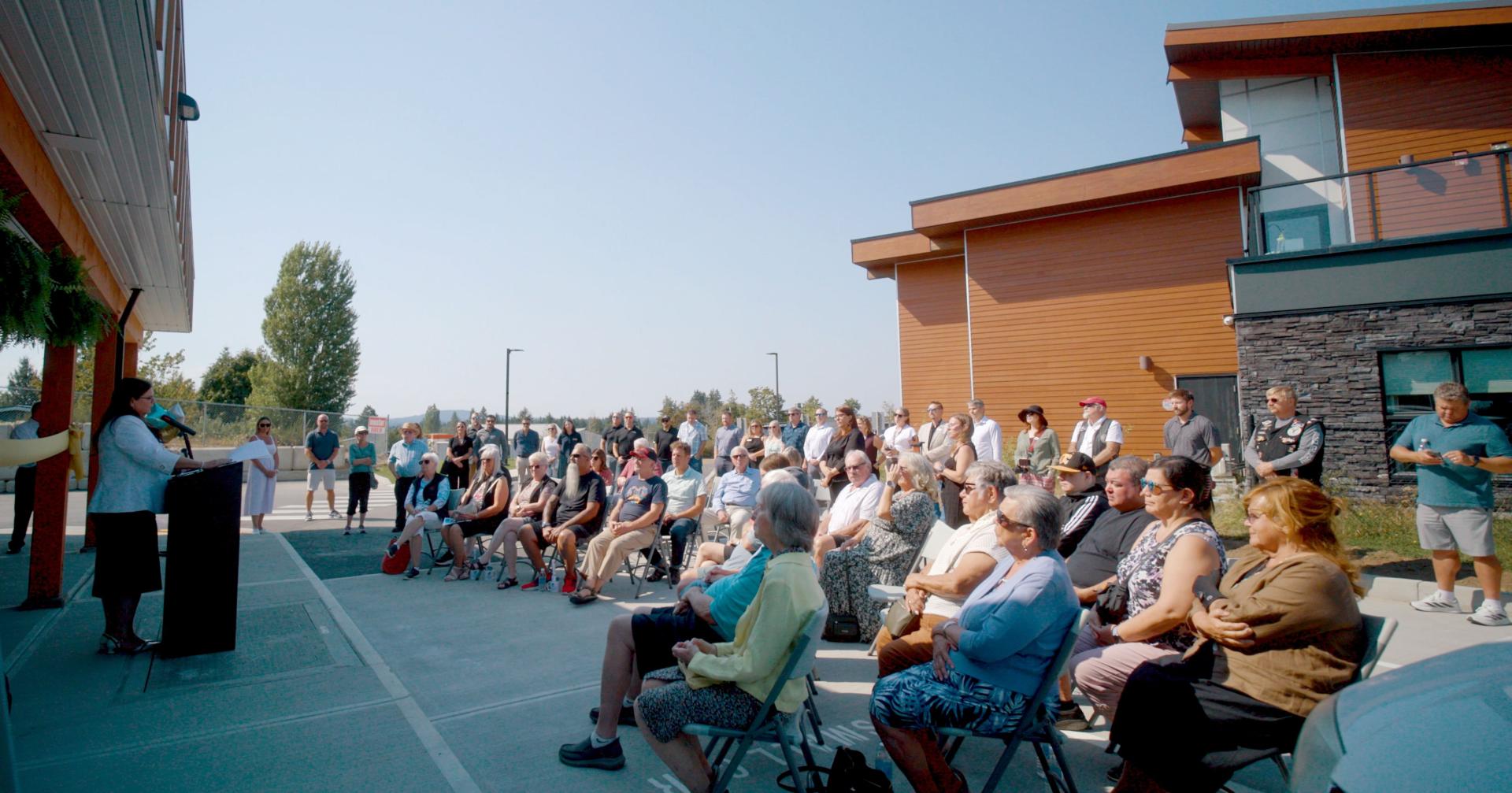 A large crowd of people seated outdoors, listening to a speaker at a podium.
